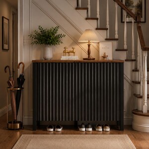 May include: A dark charcoal gray console table with a vertical ribbed texture and a wooden top. On the table sits a white vase with green foliage, two gold deer figurines, a lamp with a pleated shade, and a book with the text KINFOLK.