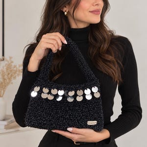 May include: A black crocheted handbag featuring a textured, shimmering yarn finish and a row of dangling, reflective silver circular sequins along the upper front. The bag includes two integrated handles and a small rectangular label at the bottom.