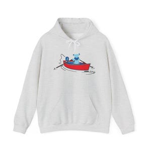 May include: A light gray heathered hooded sweatshirt featuring a graphic of a blue bear sitting in a red canoe on the water. The bear is holding a fishing rod with a blue and white circular emblem attached to the line, all centered on the chest.