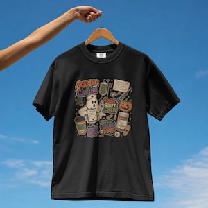Peut inclure: Un t-shirt noir à manches courtes avec une impression graphique colorée représentant un fantôme infirmier, une citrouille, du matériel médical et le texte "Scaring is Caring", "Fright Shift Crew" et "Boo-ster Shot Squad" au design rétro.
