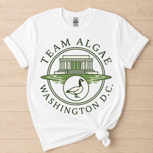 Peut inclure: Un t-shirt blanc à manches courtes avec un motif circulaire vert. Le design comprend le texte TEAM ALGAE WASHINGTON D.C., une illustration du Lincoln Memorial et un emblème circulaire central représentant une oie de profil.