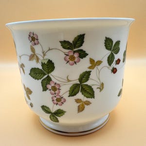 May include: A white porcelain planter featuring a botanical pattern of green leaves, small pink flowers with yellow centers, and tiny red strawberries. The vessel has a flared rim, a rounded body, and a circular base with a thin gold trim.