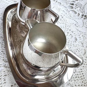 May include: A silver-toned metal creamer and sugar bowl set resting on an oval tray. The tray features an ornate embossed floral border. Both the creamer and the sugar bowl have polished, smooth surfaces, curved handles, and a reflective metallic finish.