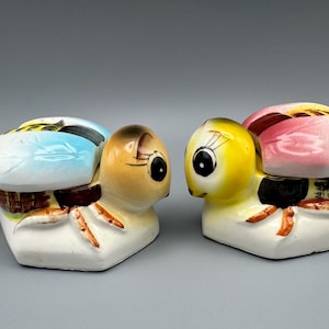May include: Two ceramic bee-shaped condiment jars with lids and small spoons. The left bee has a light blue wing and tan head, while the right bee has a pink wing and yellow head. Both feature black eyes, striped bodies, and white bases.
