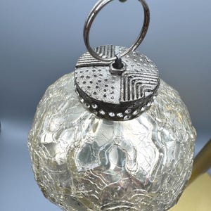 May include: A spherical glass ornament with a crackled texture and a metallic silver finish. The top features a textured silver cap with geometric patterns and a ring for hanging. The glass has a translucent, reflective quality with visible fissures.