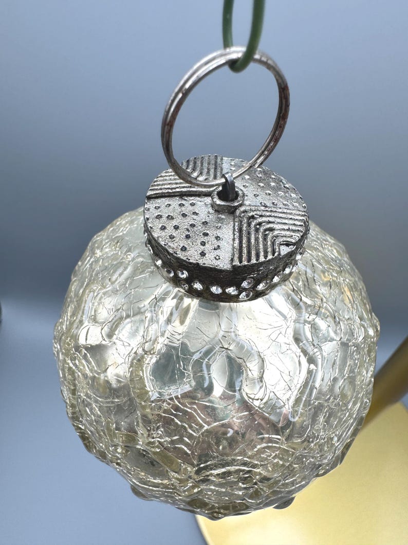 May include: A spherical glass ornament with a crackled texture and a metallic silver finish. The top features a textured silver cap with geometric patterns and a ring for hanging. The glass has a translucent, reflective quality with visible fissures.