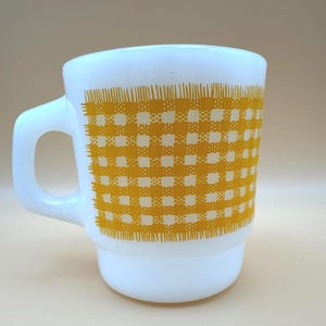 Pode incluir: Uma caneca de vidro leitoso branco com um padrão xadrez amarelo proeminente impresso na lateral. A caneca tem uma alça grossa e arredondada e uma base escalonada, com o padrão de grade amarela exibindo uma estética texturizada.