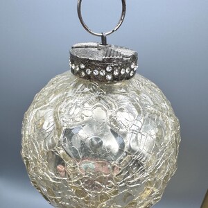 May include: A spherical glass ornament with a clear crackled texture finish. The top features a metallic cap embellished with a band of small, clear rhinestones and a circular metal hanging loop attached to the top of the decorative cap.