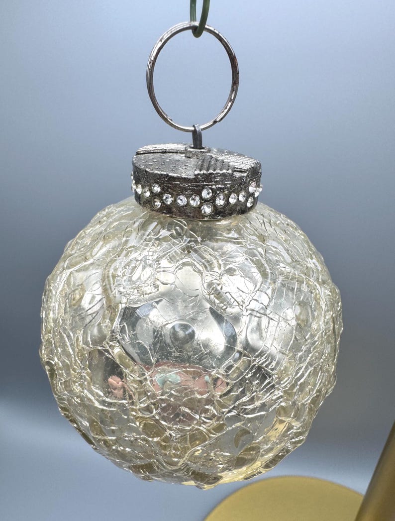 May include: A spherical glass ornament with a clear crackled texture finish. The top features a metallic cap embellished with a band of small, clear rhinestones and a circular metal hanging loop attached to the top of the decorative cap.