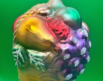 May include: A multi-colored glass ornament shaped like a basket of fruit, featuring a pear, grapes, and textured surfaces. The ornament has a gold-toned metal cap with a hanging loop and is decorated with gold glitter and white crystalline accents.