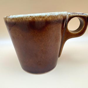 May include: A ceramic mug with a tapered, conical body featuring a glossy dark brown glaze that transitions to a lighter, creamy white drip pattern along the top rim. The mug includes a distinctive, angular handle with a circular opening.