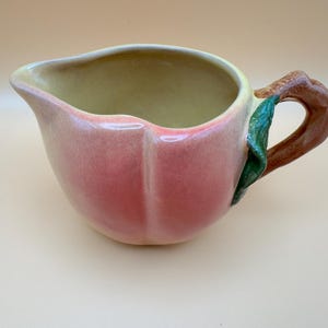 May include: A ceramic creamer pitcher shaped like a peach with a gradient pink and yellow exterior. The pitcher features a textured brown handle resembling a branch and a single green leaf attached to the side, with a smooth cream-colored interior.