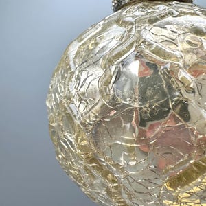 May include: A spherical glass ornament with a crackled texture and a pale yellow tint. The top features a metallic cap with a textured finish and small, clear embedded stones. The surface of the glass reflects light, highlighting the intricate web of fine lines throughout the orb.