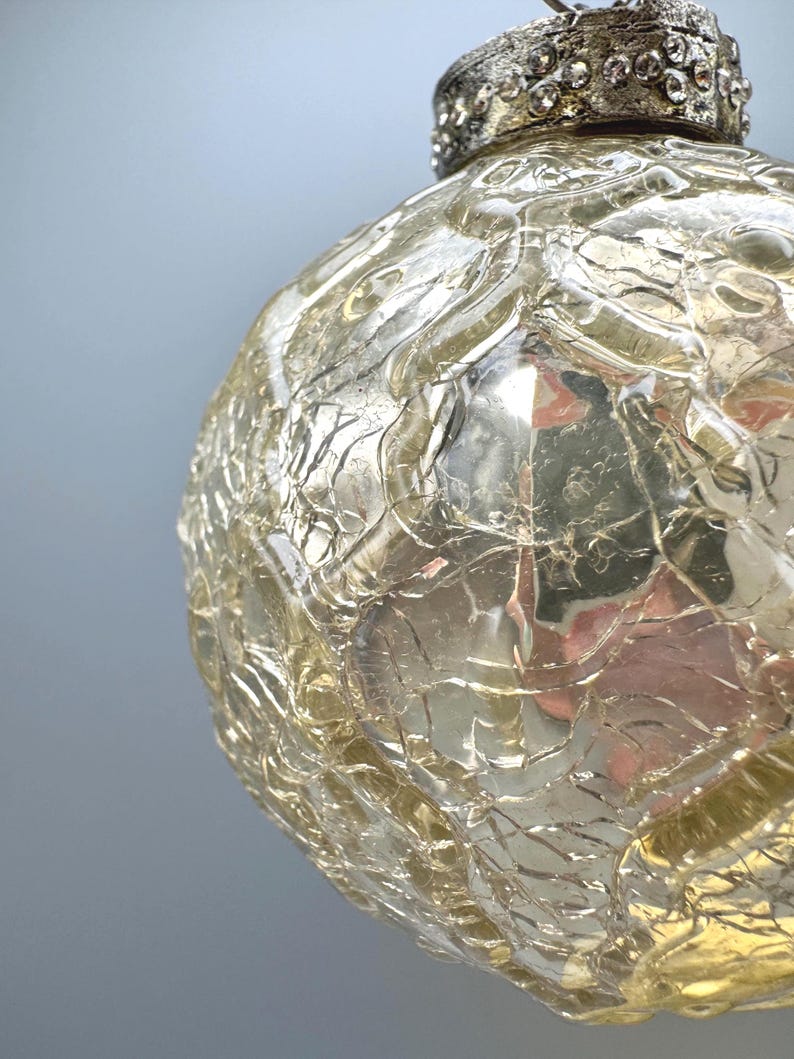 May include: A spherical glass ornament with a crackled texture and a pale yellow tint. The top features a metallic cap with a textured finish and small, clear embedded stones. The surface of the glass reflects light, highlighting the intricate web of fine lines throughout the orb.