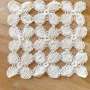 May include: A square off-white crochet doily featuring a repeating pattern of interconnected floral motifs. Each motif consists of four elongated, ribbed petals arranged in a cross shape, creating a delicate openwork design with scalloped edges.