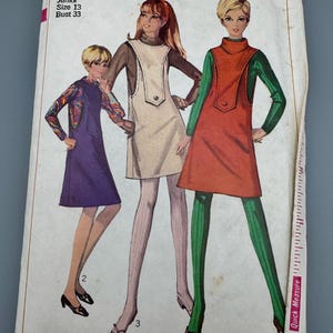Może przedstawiać: Koperta z wykrojem krawieckim vintage Simplicity 7255 z ilustracjami trzech sukienek bez rękawów o długości mini. Projekty obejmują sukienkę fioletową, kremową i pomarańczową, wszystkie noszone na bluzkach z długim rękawem.