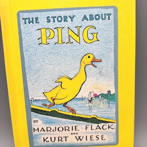 May include: A hardcover children's book titled The Story About Ping by Marjorie Flack and Kurt Wiese. The bright yellow cover features a central illustration of a yellow duck walking on a green plank over blue water with a sunset and boats.