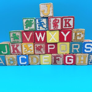 May include: A collection of vintage wooden alphabet blocks stacked in a pyramid shape. The blocks feature various letters of the alphabet, numbers, and colorful illustrations of characters including Dumbo, Pinocchio, Bambi, and Louie on a blue background.