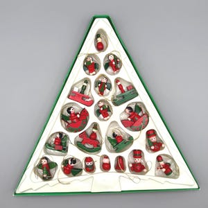 May include: A triangular box containing a set of small, hand-painted wooden Christmas ornaments. The collection includes figures of angels, rocking horses, sleds, and snowmen, all finished in festive red, green, and white paint with gold hanging strings.