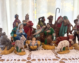 May include: A multi-piece nativity set featuring hand-painted figurines including the Holy Family, three wise men, a shepherd, a camel, a donkey, a cow, and sheep. The figures are rendered in various colors and arranged on a white lace surface.