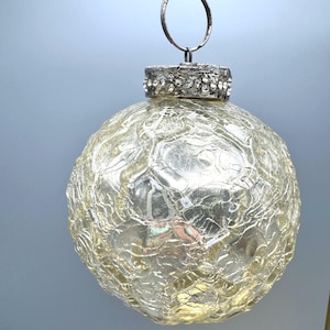 May include: A spherical glass ornament with a crackled texture and a pale gold tint. The top features a silver-toned metal cap embellished with small, clear rhinestones and a circular metal ring for hanging, providing a reflective surface.