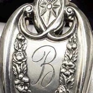 May include: A silver-toned metal spoon handle featuring an ornate embossed floral border of roses and leaves surrounding a central engraved cursive letter B. The top of the handle displays a decorative geometric star motif within a rounded frame.