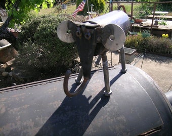 Puede incluir: Una escultura artesanal de elefante de metal con grandes orejas circulares y una trompa curvada. La escultura está hecha de piezas de metal reutilizadas y se apoya sobre cuatro patas. El elefante está colocado sobre una superficie oscura, con un fondo exterior soleado.
