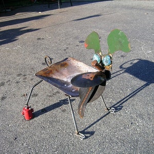 May include: A whimsical metal sculpture of a dog with green ears, blue eyes, and a rusty metal body. The sculpture is standing on a gray asphalt surface.