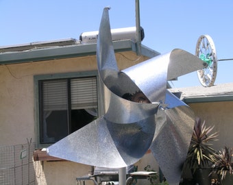 May include: A silver metal pinwheel with four blades, mounted on a pole. The pinwheel is spinning in the wind.