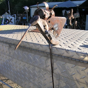 May include: A rusty metal sculpture of a bird perched on a metal surface. The sculpture is made of various metal pieces, including a wrench, a gear, and a piece of pipe. The bird's head is made of a wrench, and its beak is made of a piece of pipe. The sculpture is mounted on a metal surface that has a diamond plate pattern.