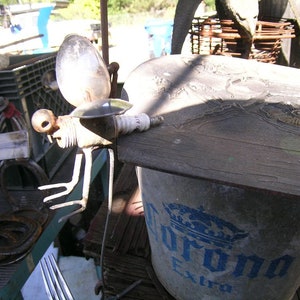 May include: A metal sculpture of a dragonfly made from recycled materials, including a light bulb, a spark plug, and metal wire. The sculpture is perched on a wooden lid of a Corona Extra beer bucket.