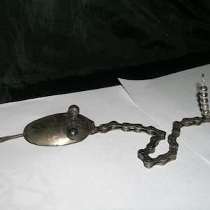 Op de afbeelding: Een zilveren lepel met een ketting eraan bevestigd, die een fantasievol ontwerp vormt. De ketting is verbonden met een kleine metalen toren gemaakt van kralen.