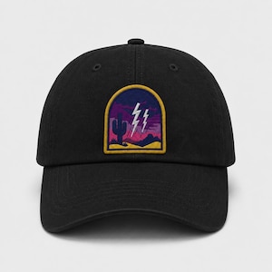 May include: A black baseball cap featuring an embroidered patch on the front. The patch is arched and displays a desert landscape with a cactus, rolling hills, and two white lightning bolts against a purple and pink sky, framed in yellow.