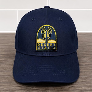 May include: A navy blue baseball cap featuring a rectangular patch with rounded top corners. The patch displays a yellow embroidered cactus graphic with radio wave symbols above it, set against a desert landscape, with the text DESERT STATIC.