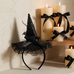 May include: A black witch hat headband featuring a pointed crown with a textured pattern, a black rose accent, and a wide brim edged with intricate black lace. The hat is adorned with a black ribbon and small feathers, creating a dark aesthetic.