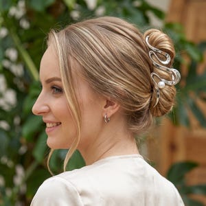 Puede incluir: Una pinza de pelo de metal plateado con un diseño fluido, abstracto y ondulado, que presenta una cuenta esférica blanca como acento. El accesorio está sujeto en un moño de cabello rubio, mostrando su superficie pulida, reflectante y su forma curva.