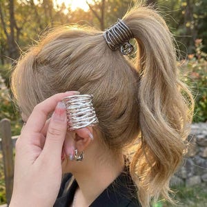 Puede incluir: Un manguito para el cabello de metal plateado con un diseño de alambre texturizado y en capas. El accesorio cilíndrico presenta un patrón abierto y entrecruzado, mostrado tanto sujeto en una coleta como sostenido por separado al frente.