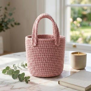 May include: A dusty pink crochet basket with a cylindrical shape and two sturdy, upright handles. The basket features a consistent, tight stitch pattern throughout its body and is constructed from a thick, durable yarn material.