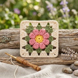 May include: A square crochet granny square featuring a central yellow flower with pink petals, surrounded by green leaves and an off-white border. A wooden crochet hook with a light-colored yarn tail is positioned in the foreground.