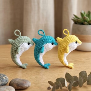 Può includere: Tre piccoli delfini all'uncinetto nei colori verde salvia, turchese brillante e giallo sole. Ogni delfino presenta una pancia bianca, un piccolo occhiello sulla parte superiore per appenderlo e occhi a perla neri, realizzati a maglia.