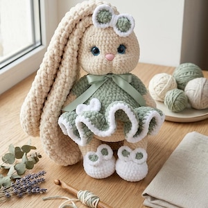 May include: A handmade crochet rabbit doll with cream-colored yarn, featuring long ears, blue eyes, and a pink nose. The rabbit wears a sage green dress with a white ruffled hem, a matching bow on its head, and white shoes with green accents.