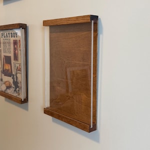 Peut inclure: Un présentoir de magazine en bois avec une façade en acrylique transparent. Le boîtier est en bois teinté foncé et est monté sur un mur blanc. Le magazine est visible à l'intérieur du boîtier.