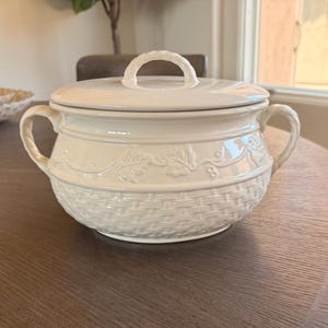 May include: A cream-colored ceramic tureen with a matching lid, featuring a woven basket-weave texture on the lower body and a raised embossed vine and leaf pattern around the upper rim, complete with two twisted rope-style side handles.