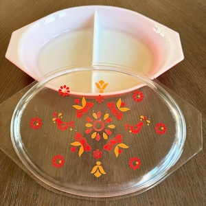 May include: A light pink divided serving dish with a matching clear glass lid. The lid features a central floral pattern in orange and red, surrounded by small red flowers and stylized bird motifs, with integrated handles on both sides.