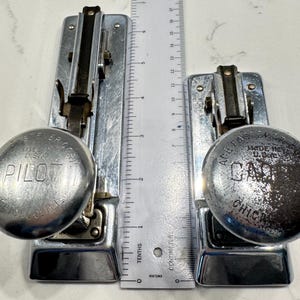 May include: Two vintage chrome staplers, one labeled Pilot and one labeled Cadet, both made in the USA in Chicago, Illinois. A white ruler is positioned between them, showing the Pilot stapler is approximately 7 inches long and the Cadet is 5 inches.