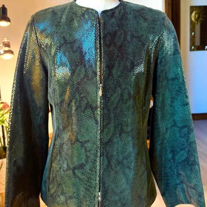 May include: A long-sleeved jacket featuring a dark green and black snakeskin-like textured pattern. The garment has a round neckline, a full-length front zipper closure with a silver-toned pull, and a structured fit displayed on a mannequin.