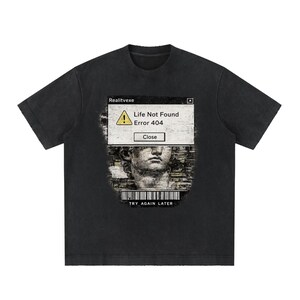 Op de afbeelding: Een zwart t-shirt met korte mouwen en een grafische print van een computervenfoutvenster met de tekst Realitvexe, Life Not Found, Error 404, Close en TRY AGAIN LATER, samen met een klassieke standbeeld-buste en een streepjescode.