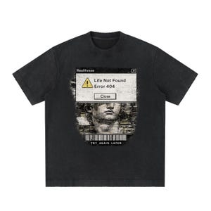 Op de afbeelding: Een zwart t-shirt met korte mouwen en een grafische print van een computervenfoutvenster met de tekst Realitvexe, Life Not Found, Error 404, Close en TRY AGAIN LATER, samen met een klassieke standbeeld-buste en een streepjescode.
