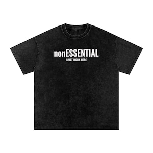 May include: A black acid-wash t-shirt with a crew neckline and short sleeves. The chest features white printed text that reads nonESSENTIAL in a bold font, with the words I JUST WORK HERE printed in a smaller, simpler font directly below it.