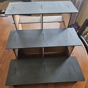May include: Three-tiered black wooden shelf unit with three shelves and four compartments on each shelf. The shelves are made of wood and have a black finish.